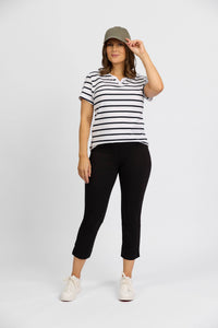 A woman stands against a white background, smiling and holding her cap as she models Vassalli’s Paris Tour Capri - Black pants, paired with a gray cap, a white and black striped short-sleeve shirt, and white sneakers.