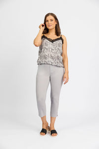 A woman with shoulder-length brown hair and a neutral expression stands against a white background, wearing a patterned sleeveless lace-trim top, Vassalli Paris Tour Capri - Silver cropped pants in stretch fabric, and black slide sandals.