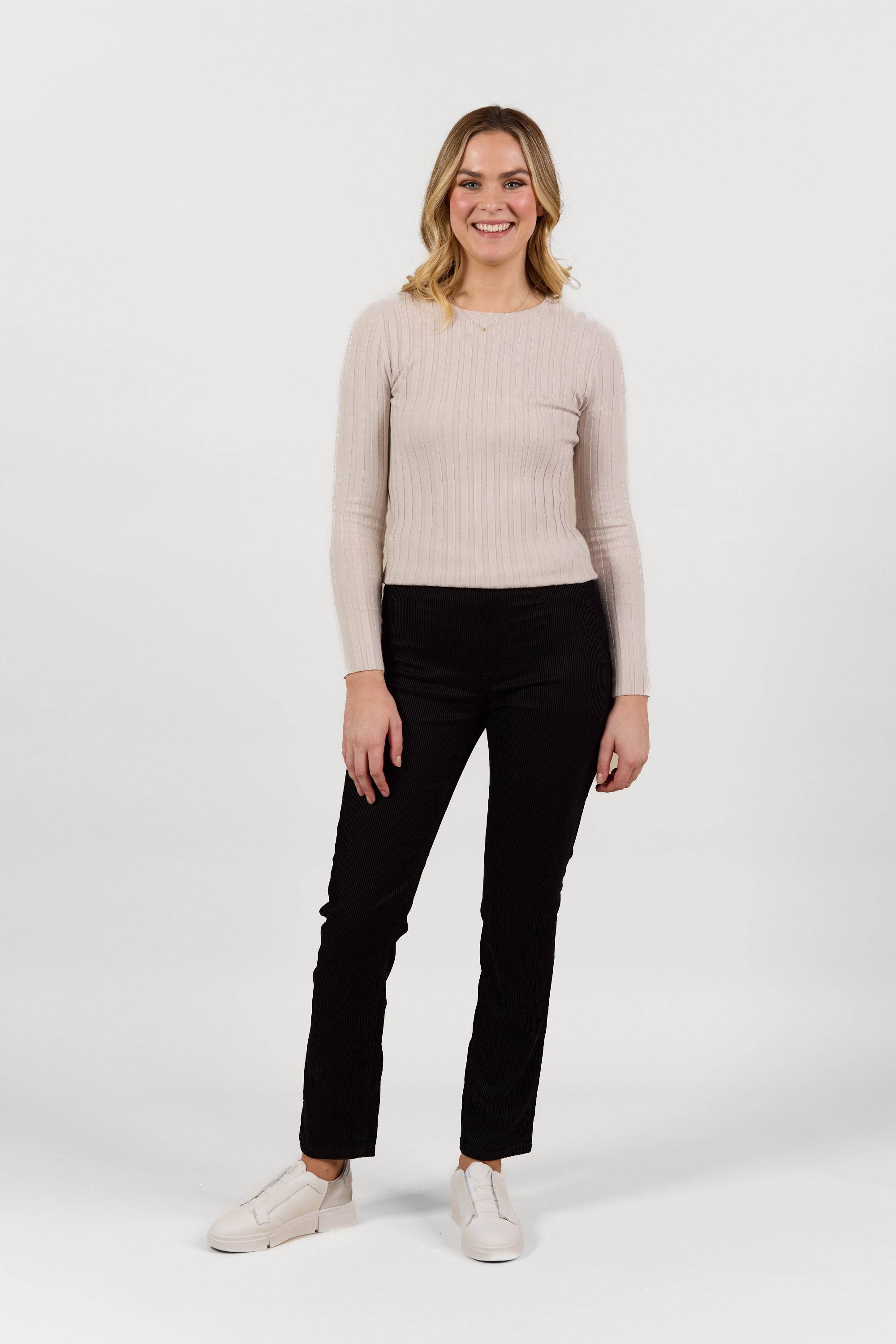 A woman stands smiling against a plain white background, wearing a long-sleeved beige ribbed top, Vassalli Bay Pull On - Black Velvet pants with an elastic waistband, and white shoes.