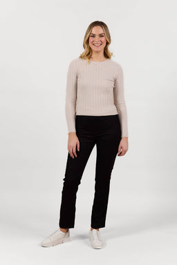 A woman stands smiling against a plain white background, wearing a long-sleeved beige ribbed top, Vassalli Bay Pull On - Black Velvet pants with an elastic waistband, and white shoes.