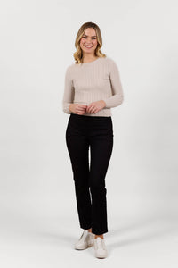 A woman with blonde hair wearing a light beige long-sleeve top, Vassalli Bay Pull On - Black Velvet pants, and white shoes stands smiling against a plain white background.
