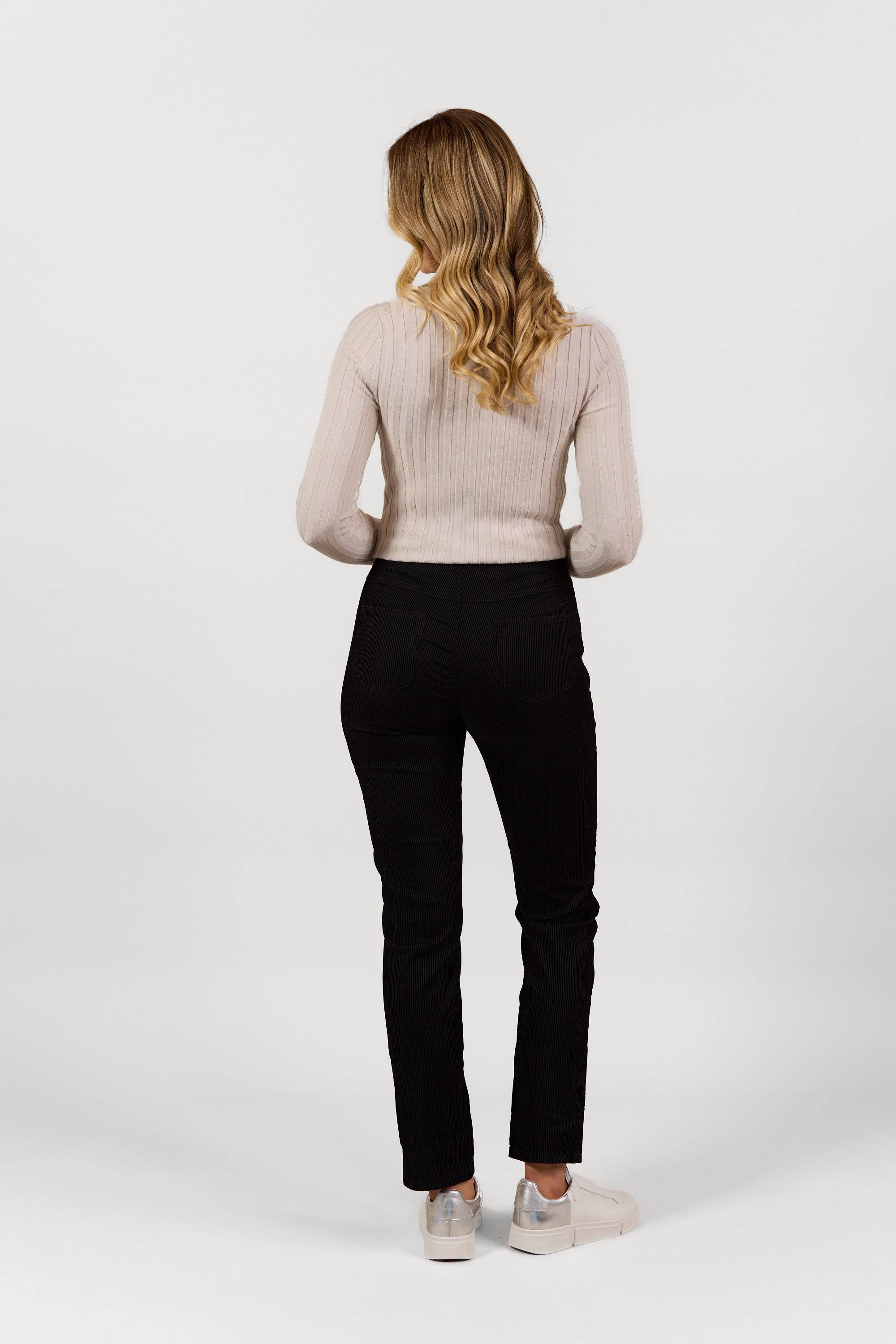A woman with wavy blonde hair, facing away from the camera, wears a light beige long-sleeve top, Vassalli Bay Pull On - Black Velvet pants, and white sneakers against a plain white background.