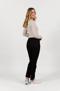 A young woman with long blonde hair, wearing a light beige long-sleeve top and Vassalli Bay Pull On - Black Velvet slim leg pants, stands sideways and smiles against a plain white background.