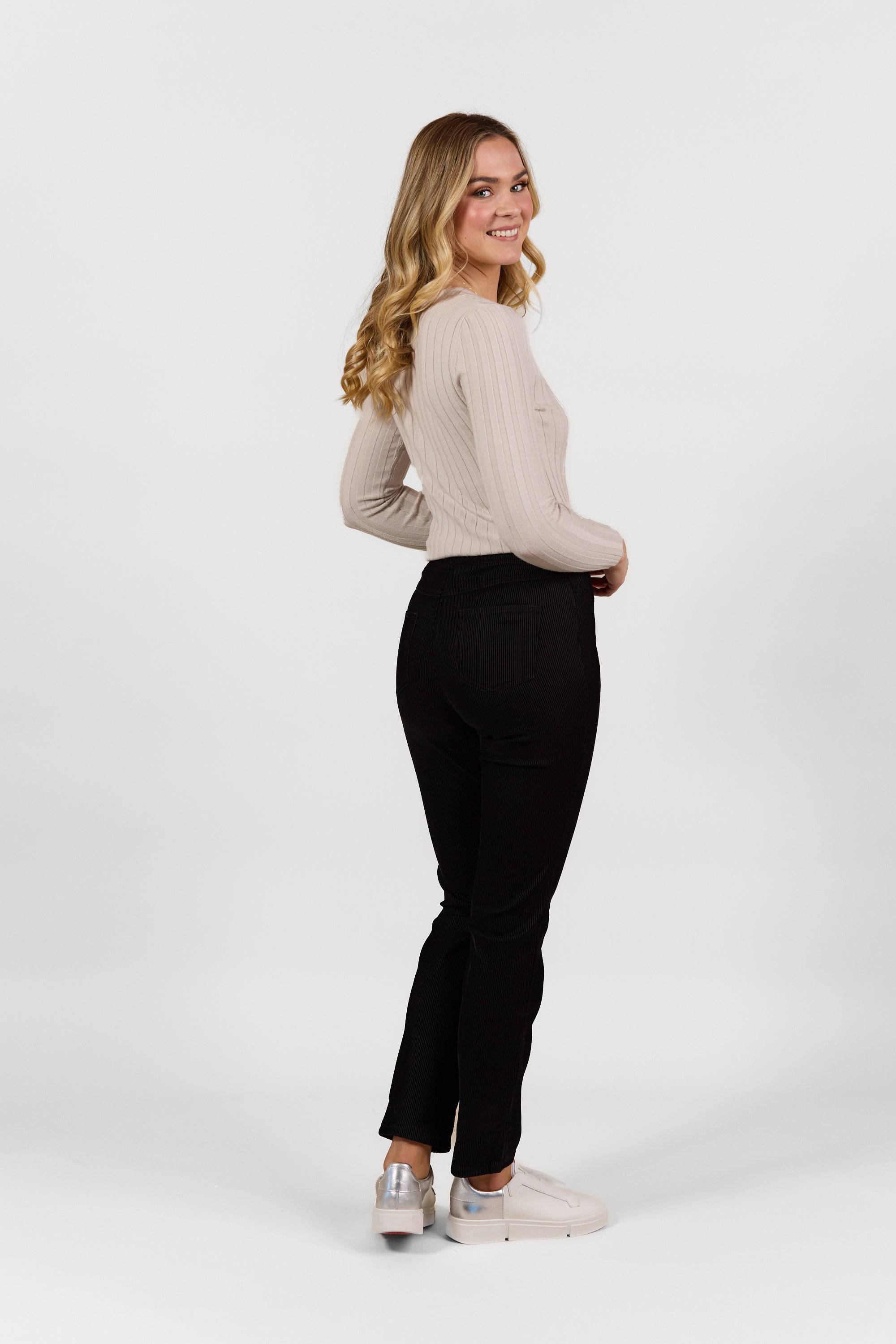 A young woman with long blonde hair, wearing a light beige long-sleeve top and Vassalli Bay Pull On - Black Velvet slim leg pants, stands sideways and smiles against a plain white background.