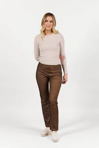 A woman with long blonde hair smiles as she poses against a white background, wearing Vassalli's Bay Pull On slim leg pants in bronze velvet and a light beige ribbed sweater with white sneakers.