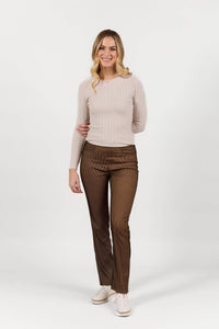 A woman with long blonde hair, wearing Vassalli Bay Pull On - Bronze Velvet slim leg pants, a light beige ribbed sweater, and white sneakers, stands against a plain white background and smiles with one arm relaxed by her side.