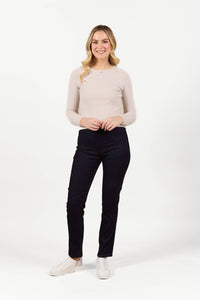 A woman with long blonde hair, wearing a light beige ribbed long-sleeve top, Bay Pull On - Navy Velvet pants by Vassalli, and white shoes, stands smiling against a plain white background.