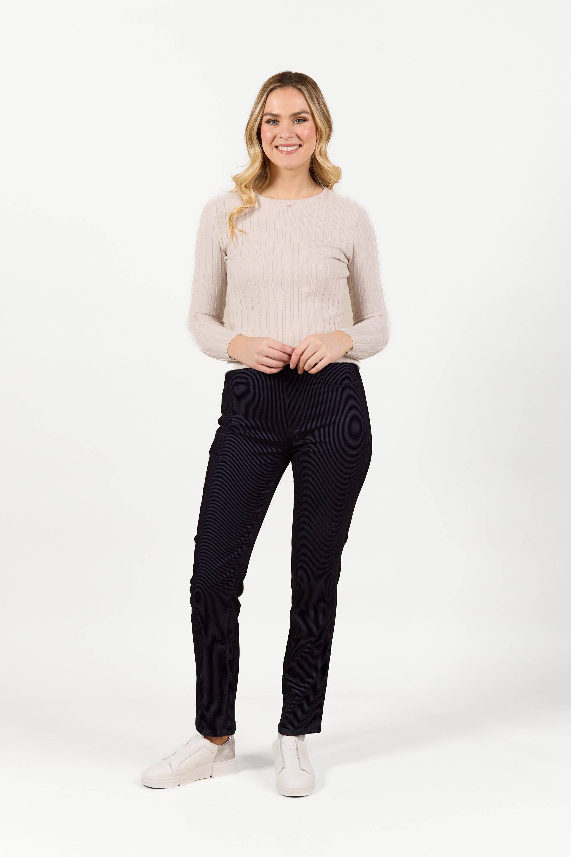 A woman with long blonde hair, wearing a light beige ribbed long-sleeve top, Bay Pull On - Navy Velvet pants by Vassalli, and white shoes, stands smiling against a plain white background.