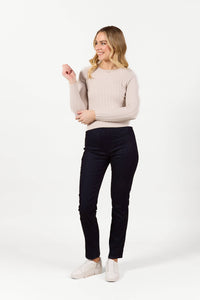 A woman smiles against a white background, wearing Vassalli's Bay Pull On slim leg pants in Navy Velvet, paired with a beige ribbed long-sleeve sweater and white slip-on shoes. She looks to the side with one arm bent.