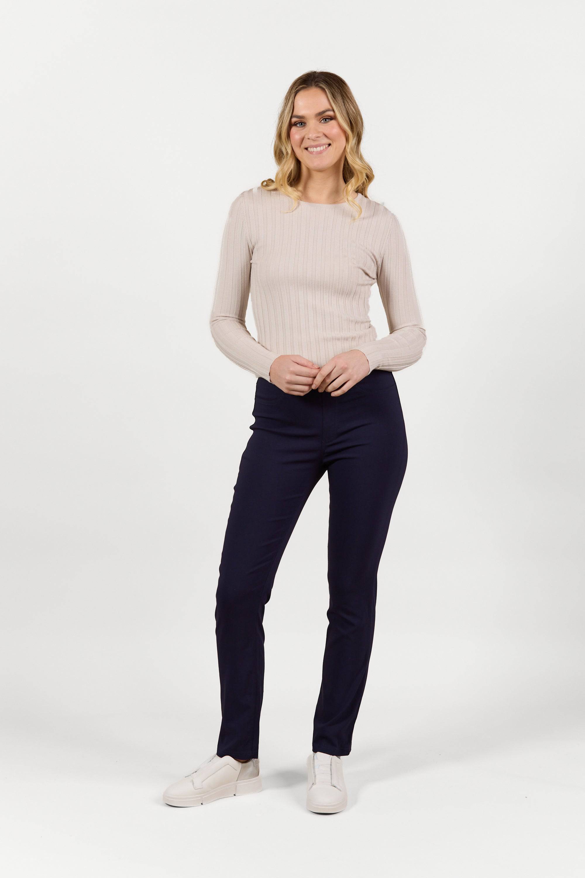 A woman stands smiling in Vassalli's Bay Pull On - Navy Bliss slim leg pants, a light beige ribbed long-sleeve top, and white slip-on shoes, posing against a plain white background.