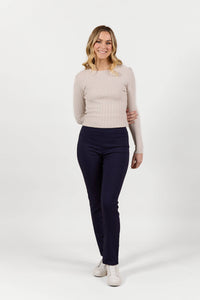 A smiling woman with blonde hair wears Vassalli Bay Pull On - Navy Bliss pants, a light beige ribbed long-sleeve top, and white shoes as she walks towards the camera against a plain white background.