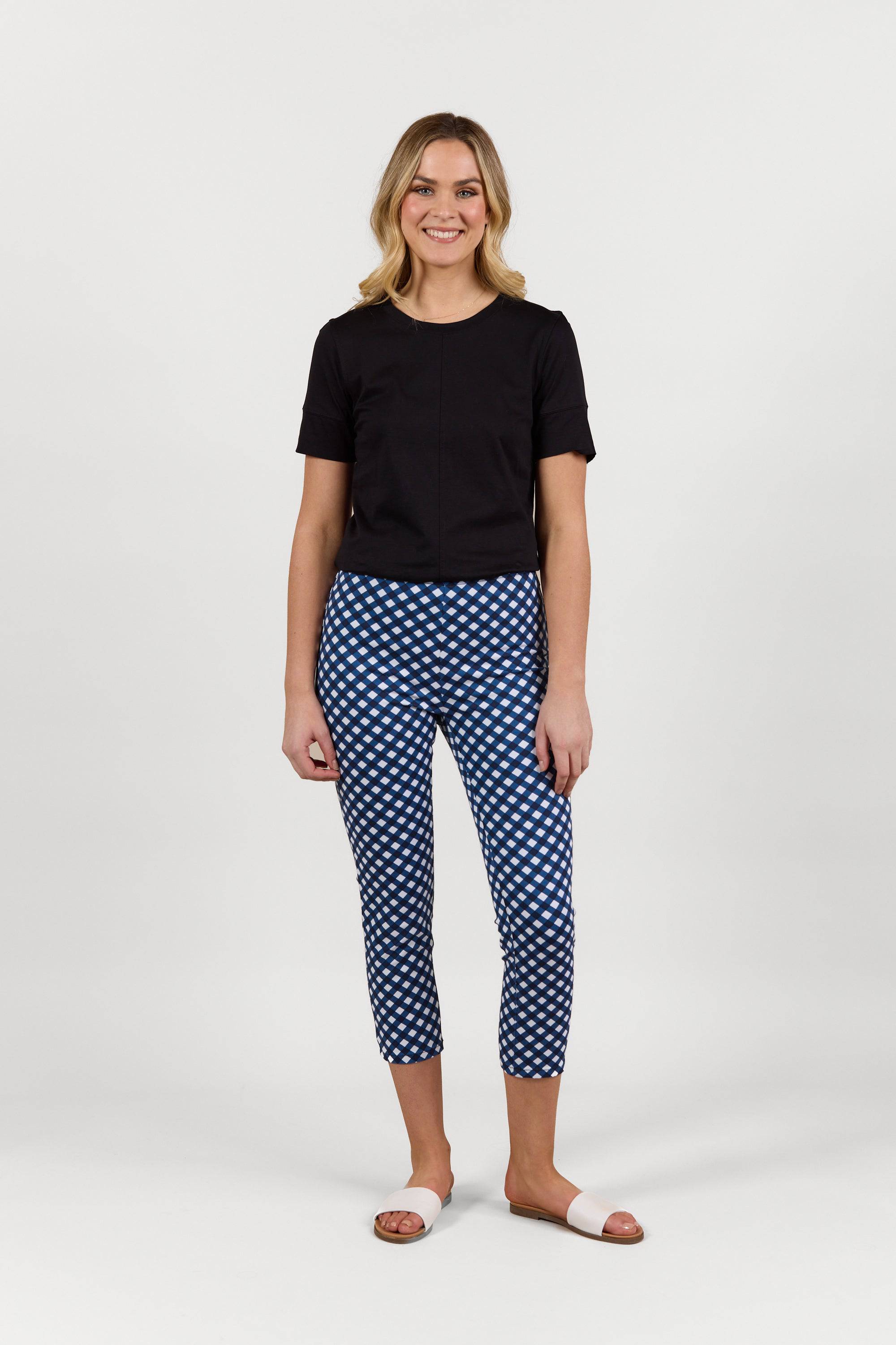 A woman smiles against a plain white background, wearing a black short-sleeve top, Vassalli's Harbour 7/8 Pant in Navy/White check, made from stretch fabric, and white slide sandals.