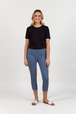 A woman smiles against a plain white background, wearing a black short-sleeve top, Vassalli's Harbour 7/8 Pant in Navy/White check, made from stretch fabric, and white slide sandals.