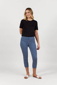 A woman smiles against a plain white background, wearing a black short-sleeve top and Vassalli Harbour 7/8 Pant in navy/white check print with stretch fabric, paired with white slide sandals.