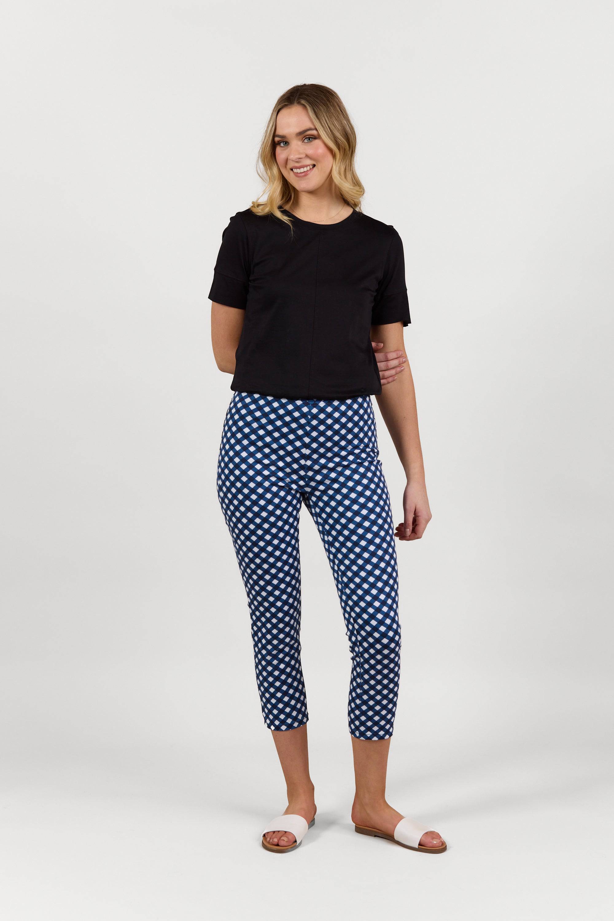 A woman smiles against a plain white background, wearing a black short-sleeve top and Vassalli Harbour 7/8 Pant in navy/white check print with stretch fabric, paired with white slide sandals.