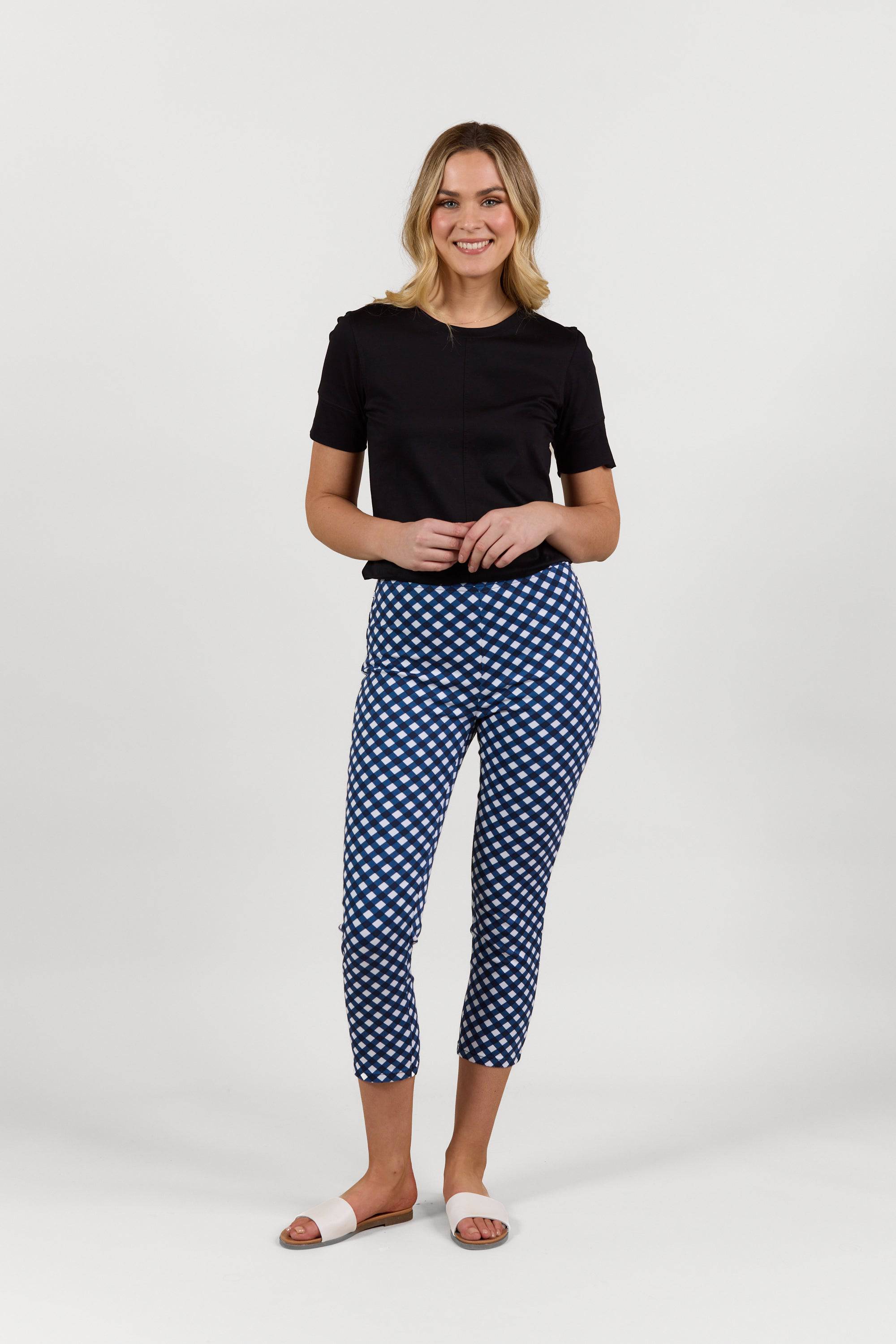 A woman smiles against a white background, wearing a black short-sleeve top, Vassalli Harbour 7/8 Pant in navy/white checkered stretch fabric, and white open-toe sandals.