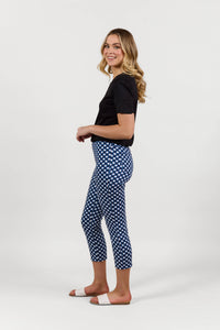 A woman with wavy blonde hair smiles sideways in a black t-shirt, Vassalli Harbour 7/8 Pant in Navy/White, and white slide sandals against a plain white background.