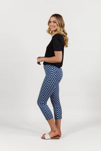 A woman stands sideways, smiling, wearing a black t-shirt, Vassalli Harbour 7/8 Pant in Navy/White with a blue-and-white check pattern, and white slide sandals against a plain white background.