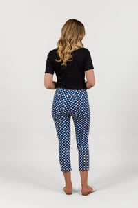 A woman with long, wavy blonde hair stands facing away, wearing a black short-sleeve top and Vassalli Harbour 7/8 Pant in navy/white checkered stretch fabric with white slip-on shoes against a plain white background.
