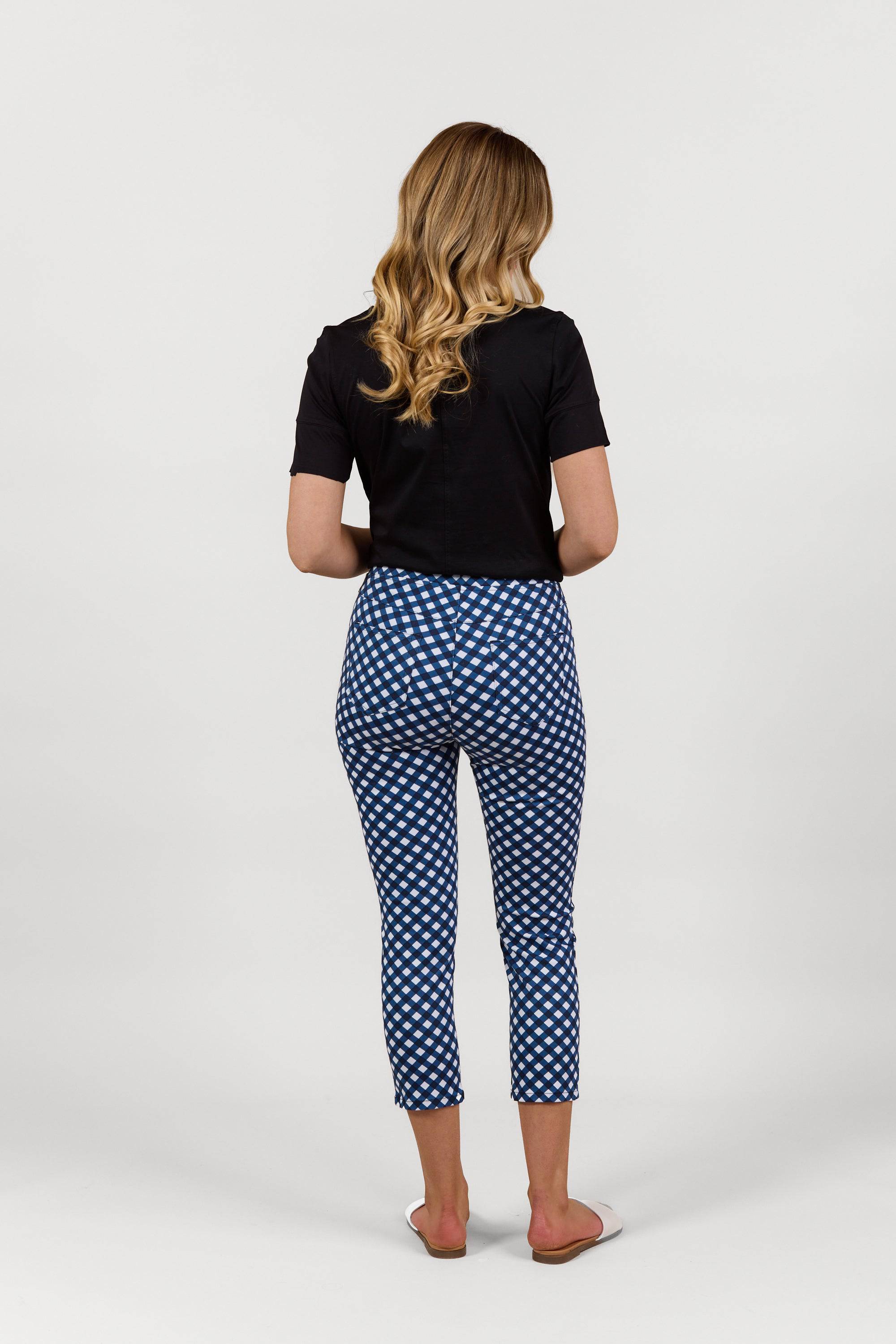 A woman with long, wavy blonde hair stands facing away, wearing a black short-sleeve top and Vassalli Harbour 7/8 Pant in navy/white checkered stretch fabric with white slip-on shoes against a plain white background.