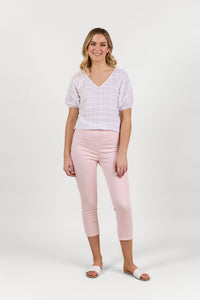 A woman stands smiling against a white background, wearing the Vassalli Harbour 7/8 Pant in Powder Pink, a white short-sleeve blouse, and white slide sandals.
