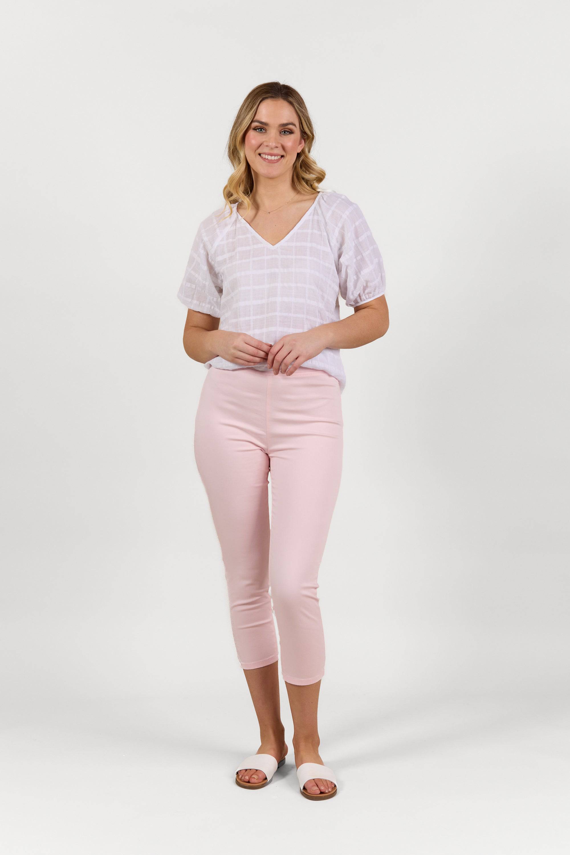 A smiling blonde woman stands against a plain white background wearing Vassalli's Harbour 7/8 Pant in Powder Pink, a white checkered short-sleeve blouse, and white slide sandals.
