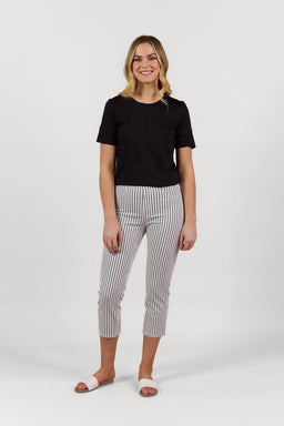 A person smiles against a plain white background, wearing a black short-sleeve top, Vassalli Sierra 7/8 Pant - Black Stripe/White, and white slide sandals.