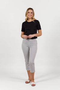 A woman smiles against a white background, wearing a black short-sleeve top, Vassalli Sierra 7/8 Pant in Black Stripe/White, and white slide sandals.