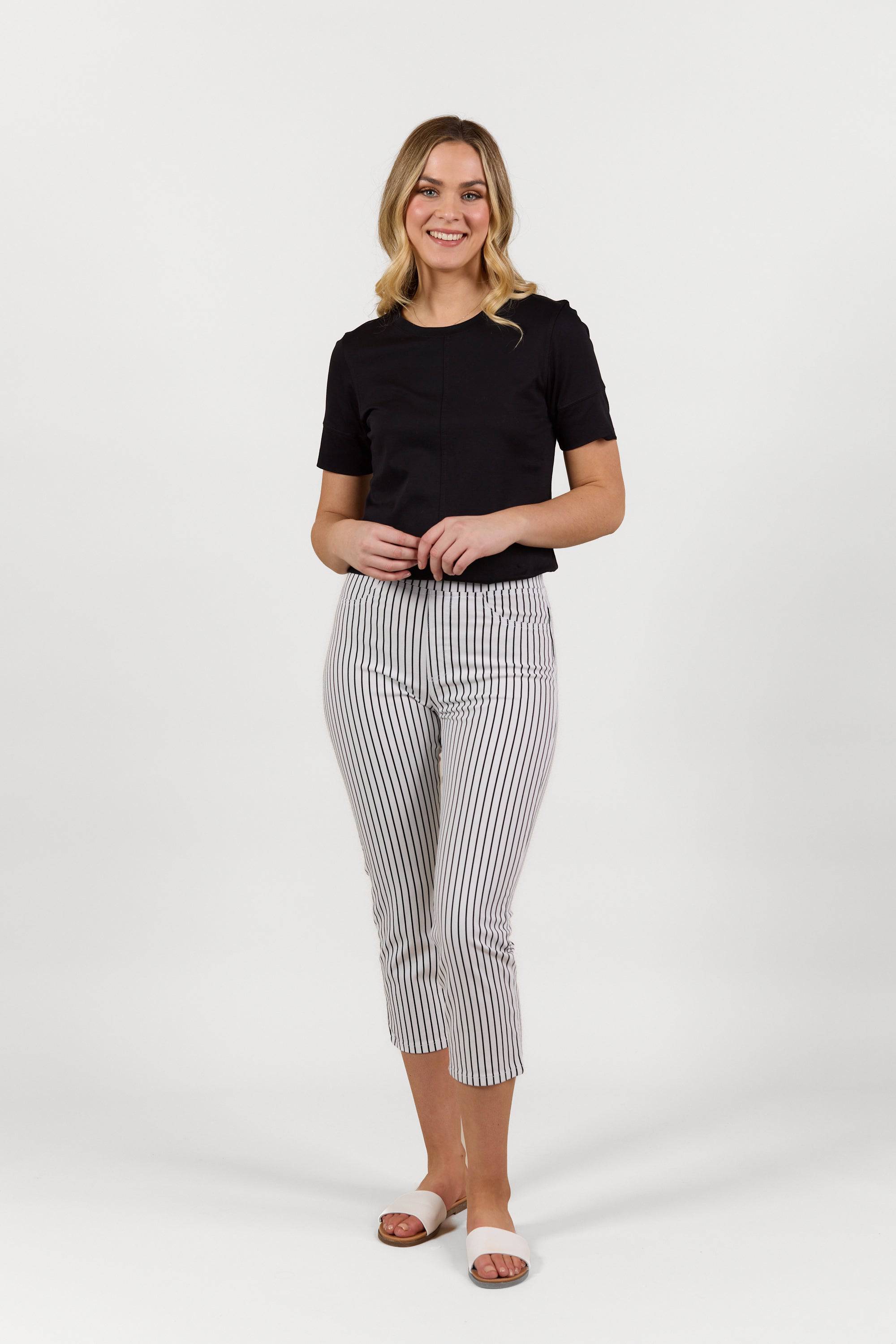 A woman smiles against a white background, wearing a black short-sleeve top, Vassalli Sierra 7/8 Pant in Black Stripe/White, and white slide sandals.