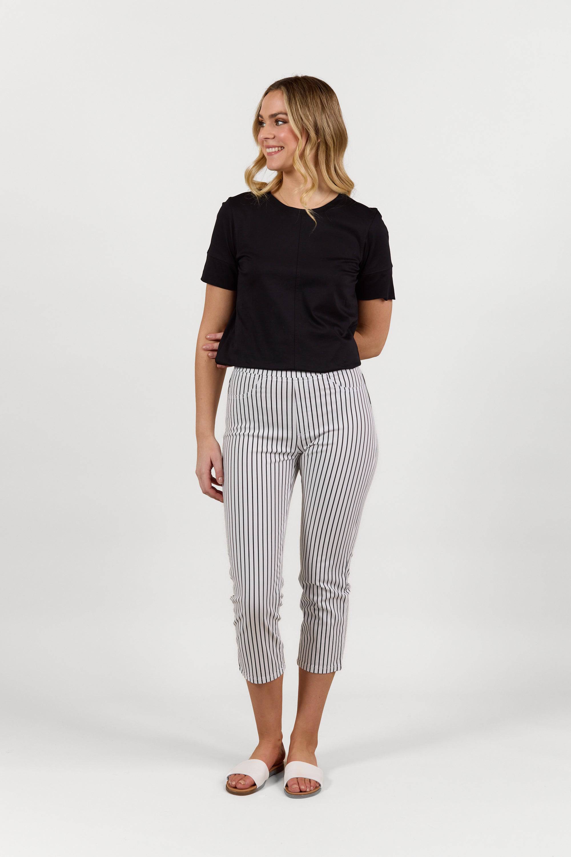 A woman with wavy blonde hair smiles, wearing Vassalli’s Sierra 7/8 Pant in Black Stripe/White, a black short-sleeve top, and white sandals, posing against a plain white background.