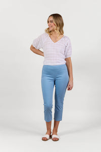 A woman smiles, wearing a sheer white short-sleeve top, Vassalli Sierra 7/8 Pant in Fresh Blue, and white slide sandals, against a plain white background.