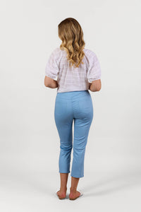 A woman with wavy blonde hair stands facing away, wearing a white short-sleeve blouse and Vassalli Sierra 7/8 Pant in Fresh Blue, styled with white flat sandals against a plain white background.