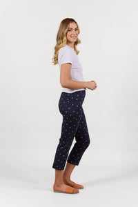 A woman with long blonde hair, wearing a white short-sleeve top and Vassalli's Sierra 7/8 Pant - Navy Daisy, stands sideways in orange sandals and smiles against a plain white background.