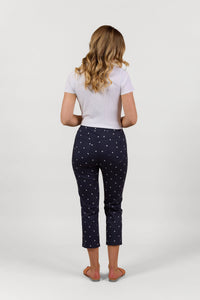 A woman with wavy blonde hair faces away, wearing a white short-sleeve top and Vassalli Sierra 7/8 Pant - Navy Daisy with a small white print and sandals, against a plain white background.