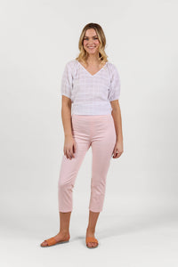 A woman smiles against a white background, wearing a white short-sleeve blouse, Vassalli Sierra 7/8 Pant in Powder Pink, and brown slide sandals.