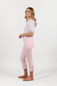 A woman with long blonde hair, wearing a white blouse and Vassalli's Sierra 7/8 Pant in Powder Pink, stands sideways and smiles against a plain white background.