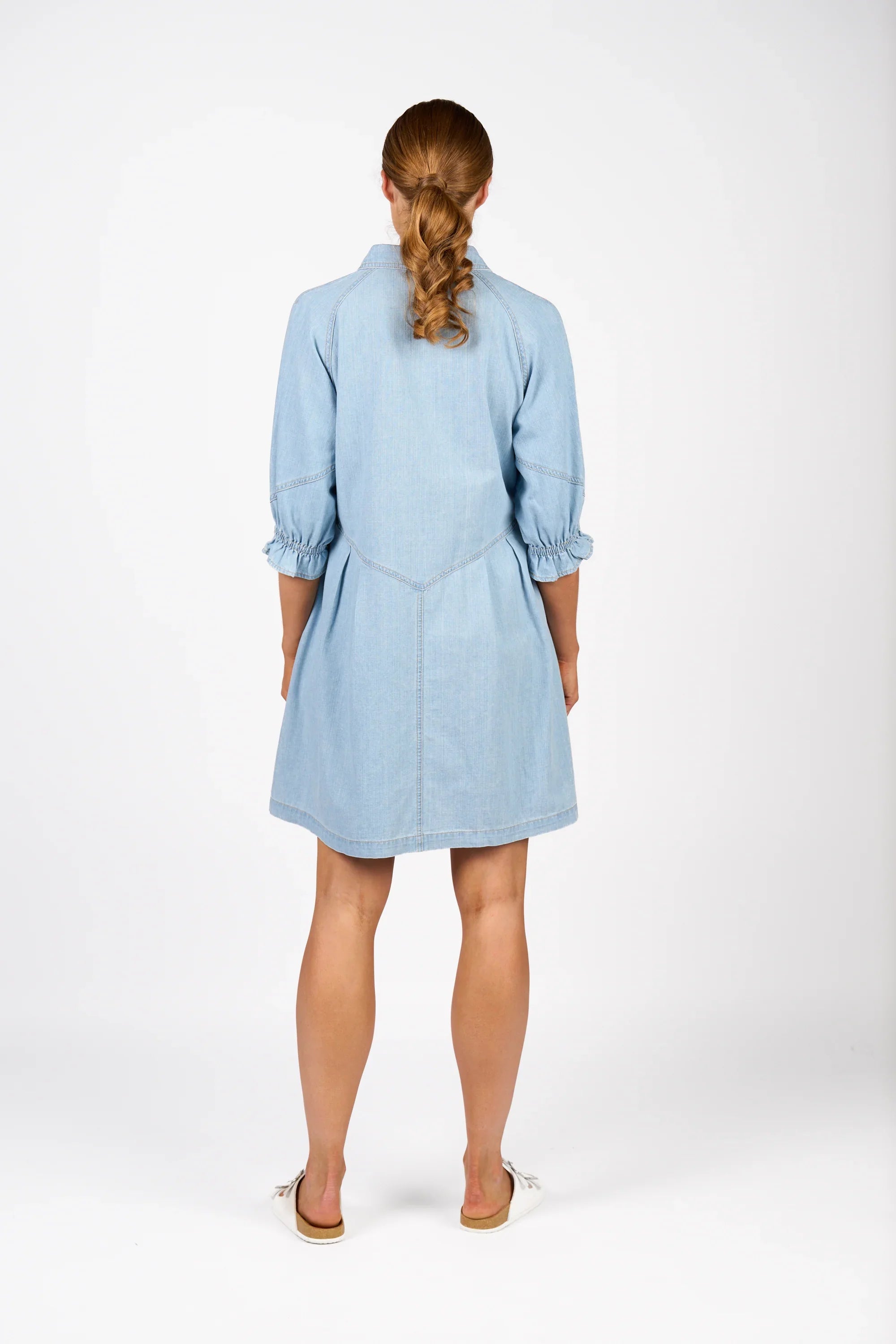 Back view of woman wearing light blue denim dress with puff sleeves, white flat shoes