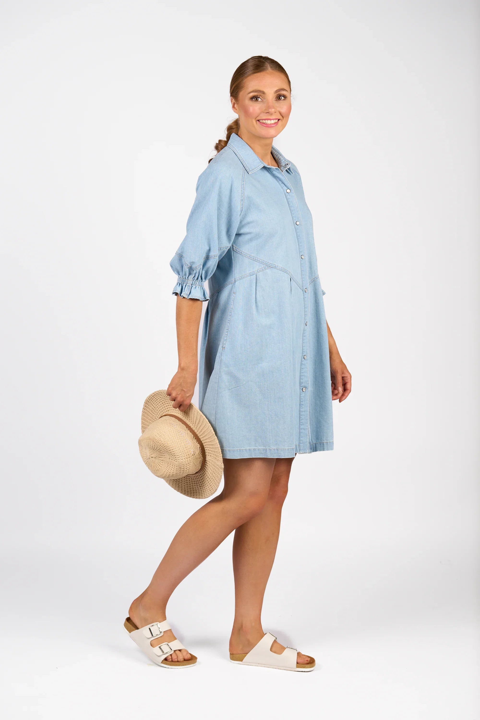 Woman in light wash denim shirt dress with ruffle sleeves, holding straw hat, white sandals