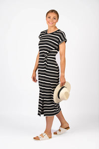 Woman in black and white striped midi dress holding straw hat, wearing white sandals, standing against a white background.