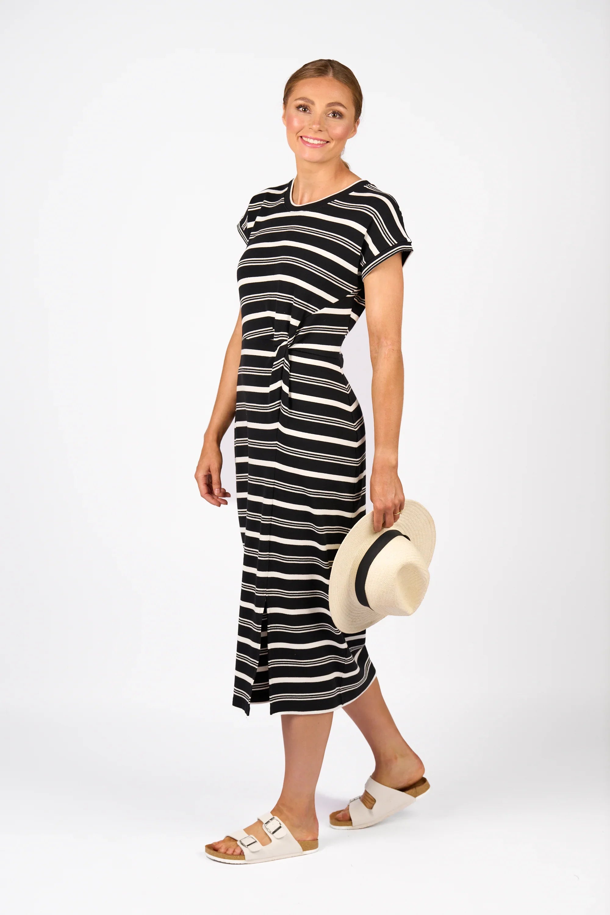 Woman in black and white striped midi dress holding straw hat, wearing white sandals, standing against a white background.