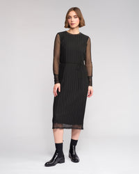 A woman poses against a plain background in the Knewe Label Freddie Dress - Black, featuring sheer long sleeves and vertical pleats, styled with black ankle boots—the ideal slim-fit winter dress for any occasion.