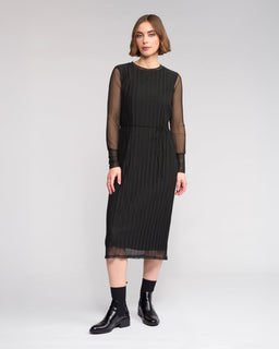 A woman poses against a plain background in the Knewe Label Freddie Dress - Black, featuring sheer long sleeves and vertical pleats, styled with black ankle boots—the ideal slim-fit winter dress for any occasion.