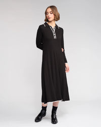 A woman stands against a light gray background wearing the Knewe Label Ritual Dress in black—a ribbed knit piece with a shirt-style collar, white trim, and black ankle boots for a chic winter look.