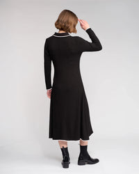 A person with short hair, seen from behind against a plain light backdrop, wears the Knewe Label Ritual Dress in black featuring a white shirt-style collar and cuffs, paired with black ankle boots.