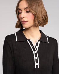 A woman with short, light brown hair looks down to her left, wearing the Knewe Label Ritual Dress in black—a ribbed knit style with a shirt collar, white trim, and white buttons. The background is light gray.