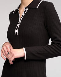 A person wears the Ritual Dress in black by Knewe Label, featuring long sleeves, ribbed knit fabric, white trim and buttons on a shirt-style collar. Their hands show several silver rings against a plain, light background.