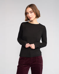 A woman with chin-length brown hair wears the Knewe Label Hero Top in black with burgundy pants, standing against a plain light gray background, hands loosely clasped.