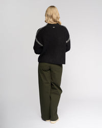 A person with blonde hair faces away, wearing the Knewe Label Lennox Sweater in Black/Winter White, paired with olive green wide-leg pants and cream shoes against a plain light background.