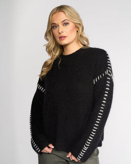 A woman with long blonde hair wears the Knewe Label Lennox Sweater in Black/Winter White, featuring white sleeve stitching, paired with dark green pants against a plain light background.