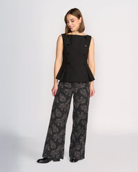A woman stands against a plain light background wearing the Knewe Label Austin Top in black, a sleeveless vest with black button detailing, paired with wide-leg dark floral print pants. She looks down and smiles, arms relaxed at her sides.